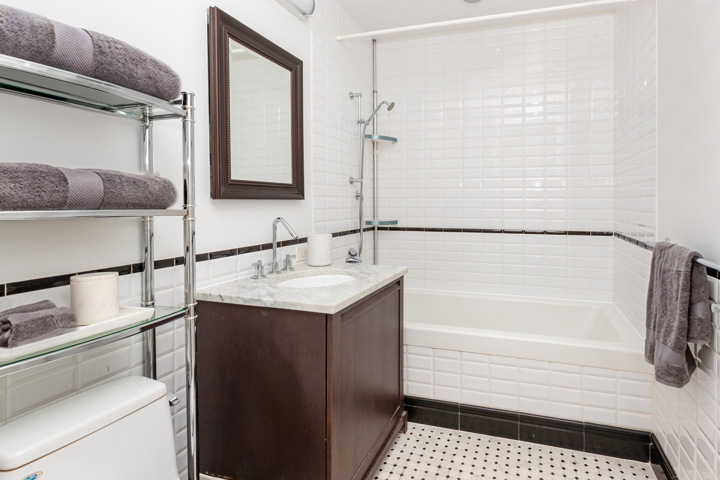 Bathroom with a white tiled bathtub, a dark wood vanity with a marble top, a mirror, and gray towels on a silver rack.