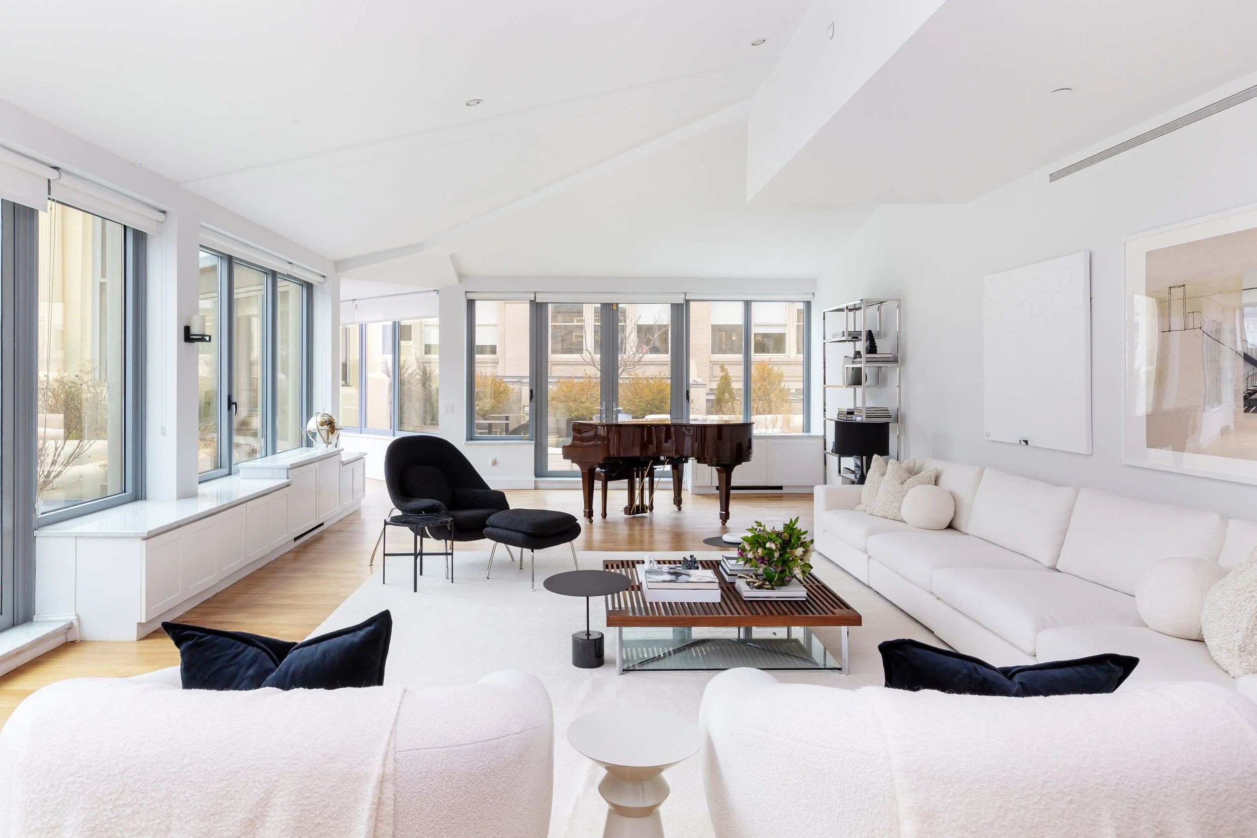 Modern living room with large windows, white walls, black chairs, a white sofa, a wooden coffee table with books and a plant, and a grand piano.