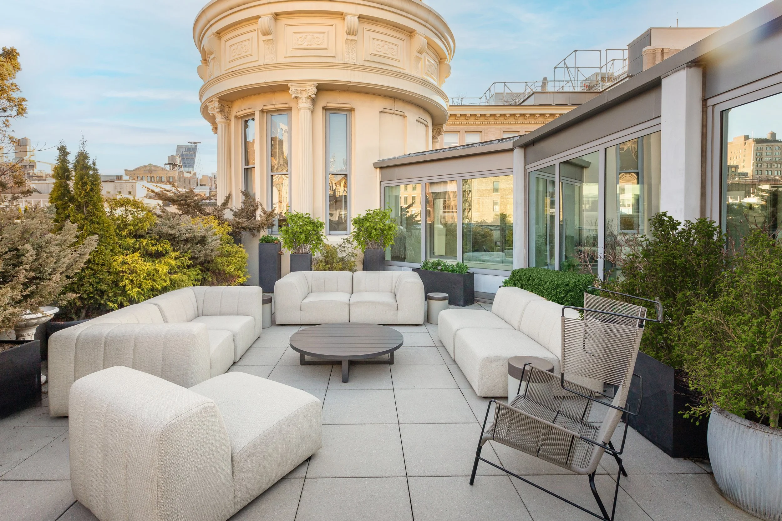 A rooftop terrace with white outdoor couches, a round black coffee table, and potted plants, overlooking a cityscape with historic and modern buildings.