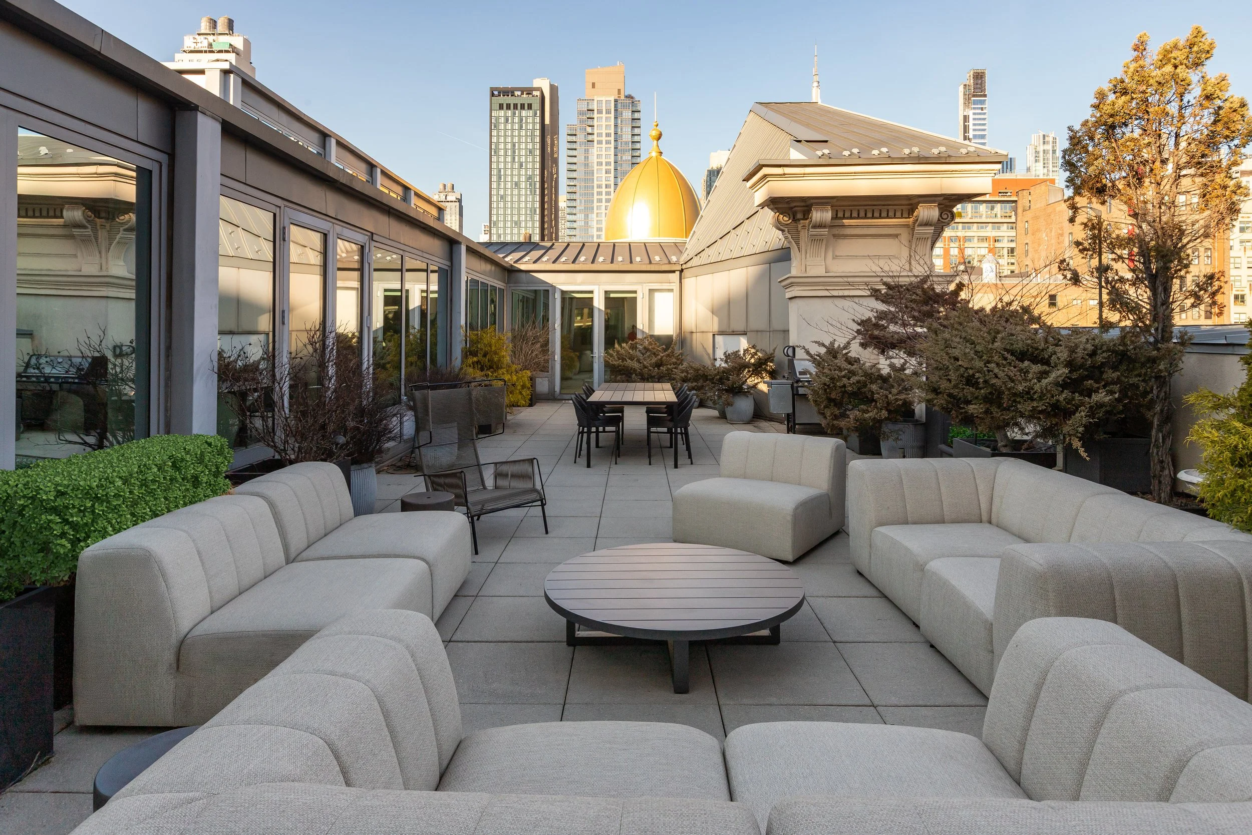 Rooftop terrace with beige sectional sofas, black chairs, a round coffee table, potted trees, city skyline with tall buildings and a golden dome in the background.