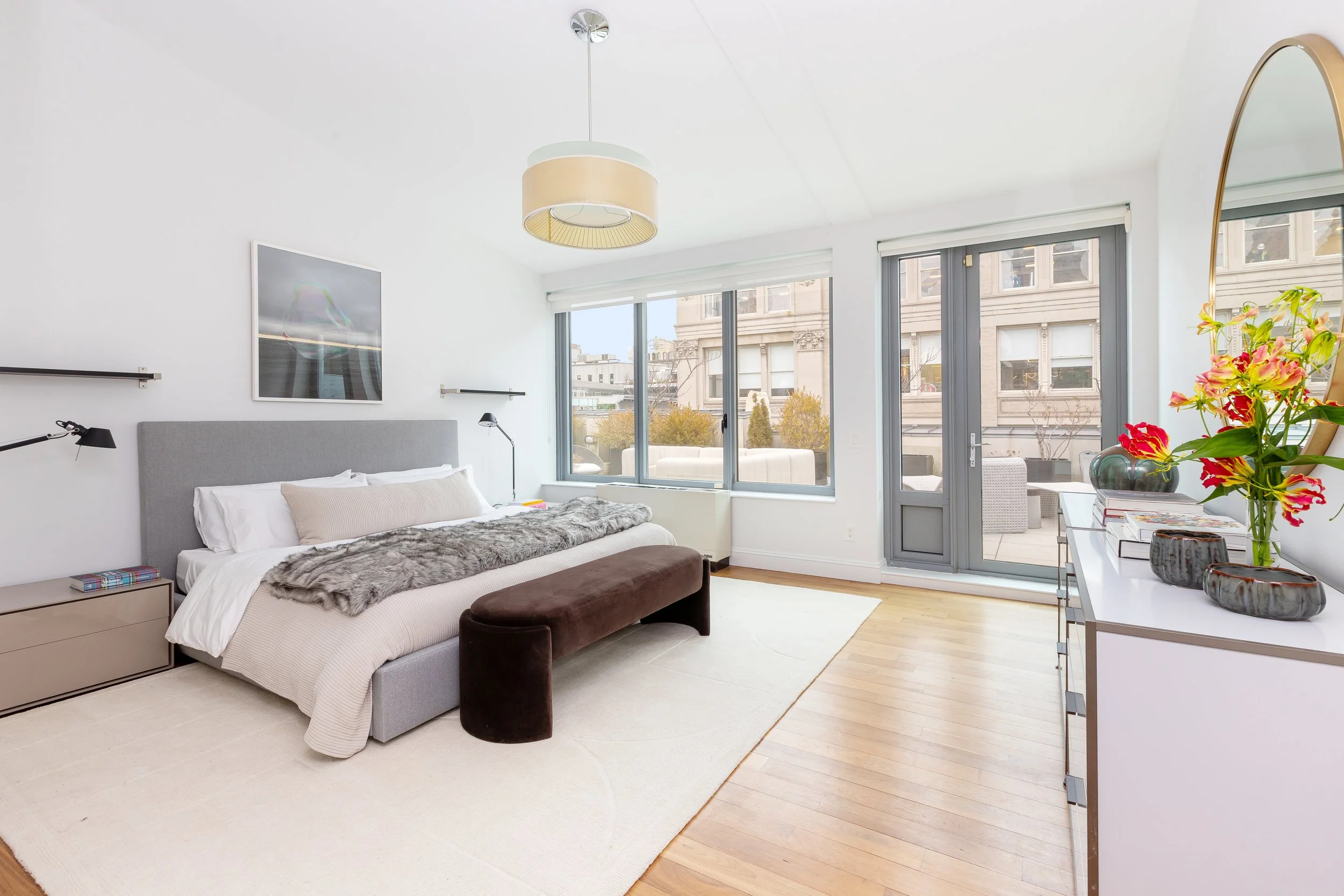 Bright bedroom with large windows, a bed with gray headboard, white bedding, and a fur throw, a brown bench at the foot of the bed, and a white dresser with flower vases and a mirror.