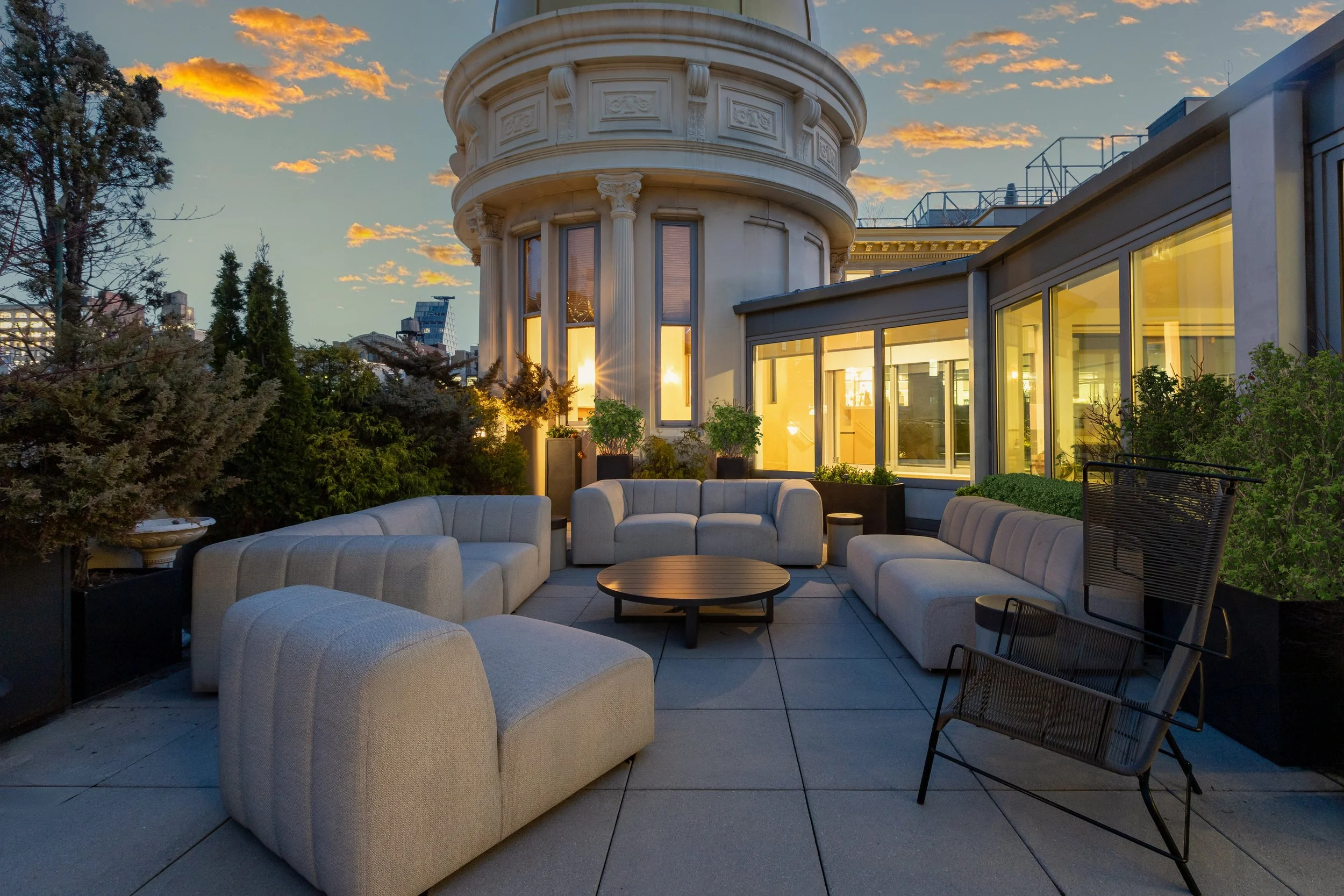 Outdoor rooftop terrace with modern beige sofas and chairs, a round coffee table, and potted plants, with a historic building with ornate architectural details in the background during sunset.
