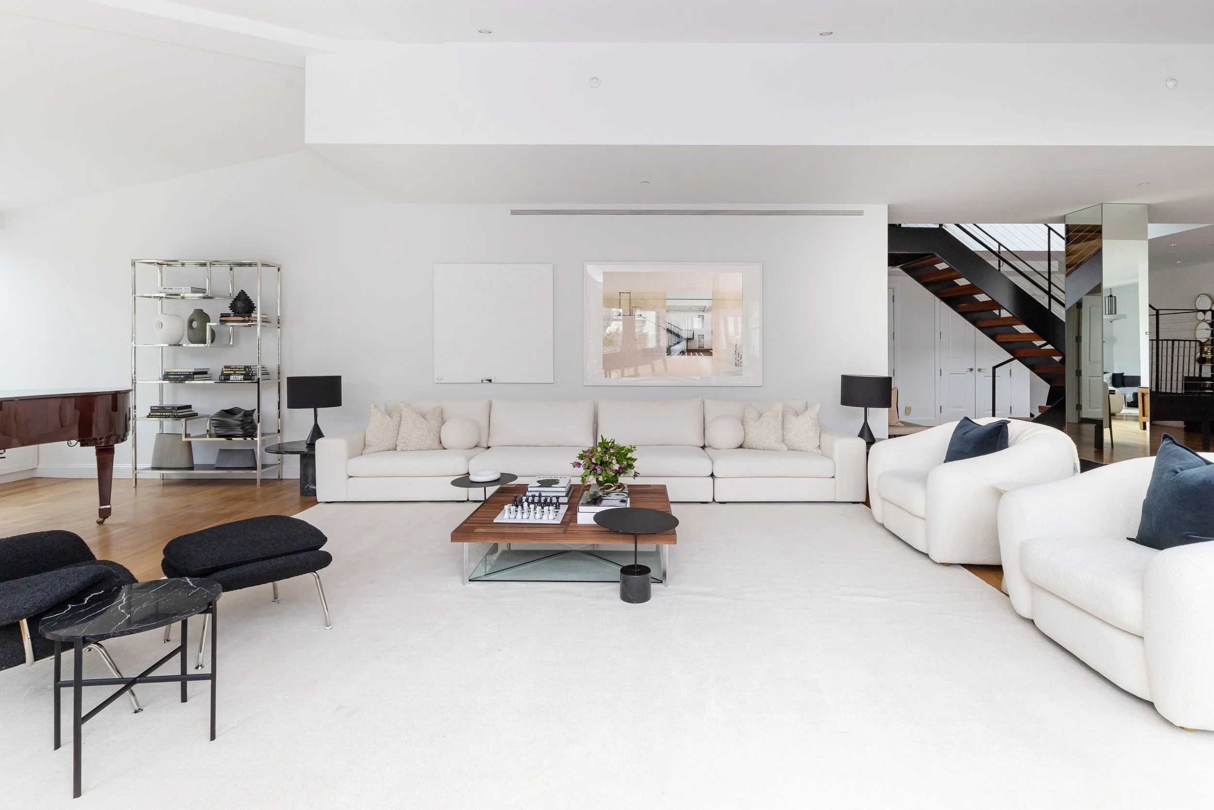 Modern living room with white sofas, a coffee table, and black lamps, featuring a white wall with framed artworks and a staircase in the background.