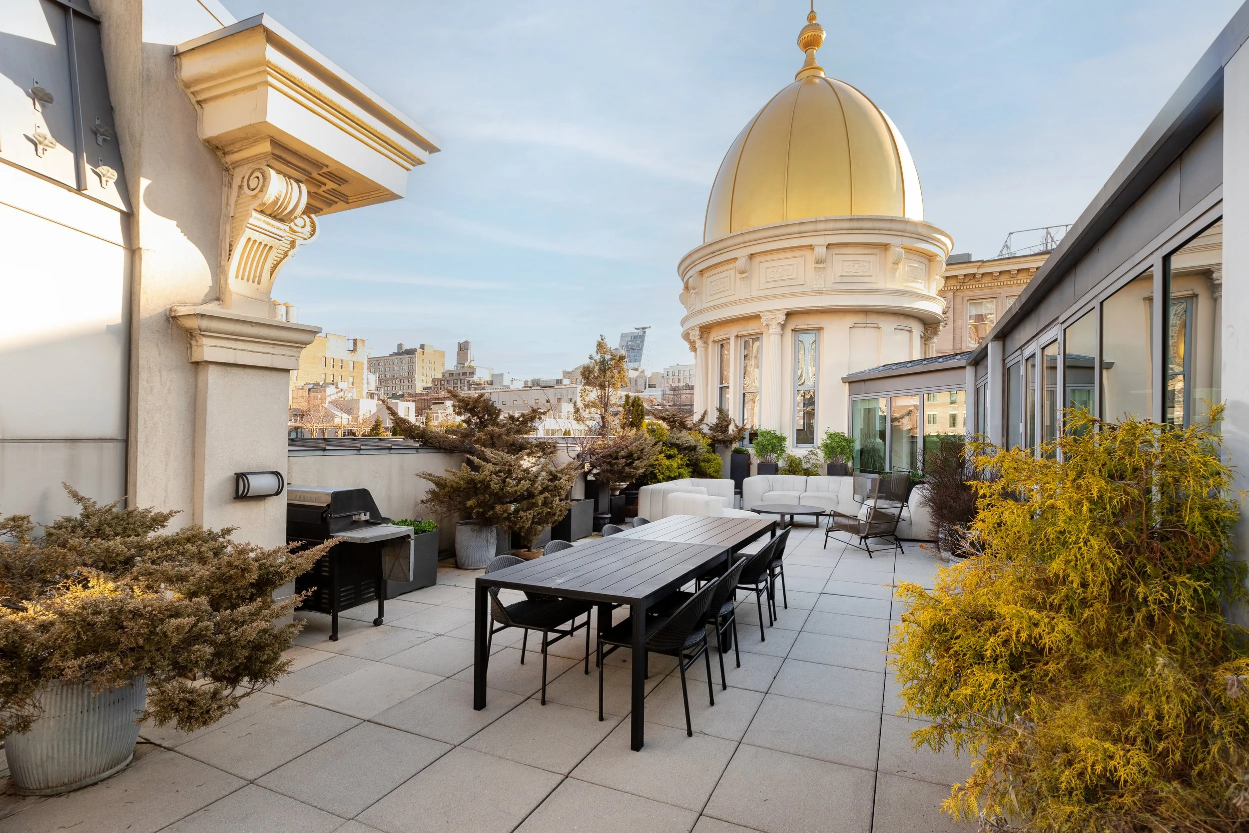 Rooftop terrace with outdoor furniture including a black dining table, chairs, sofas, and potted plants, featuring a view of a domed building with a gold roof in an urban area.