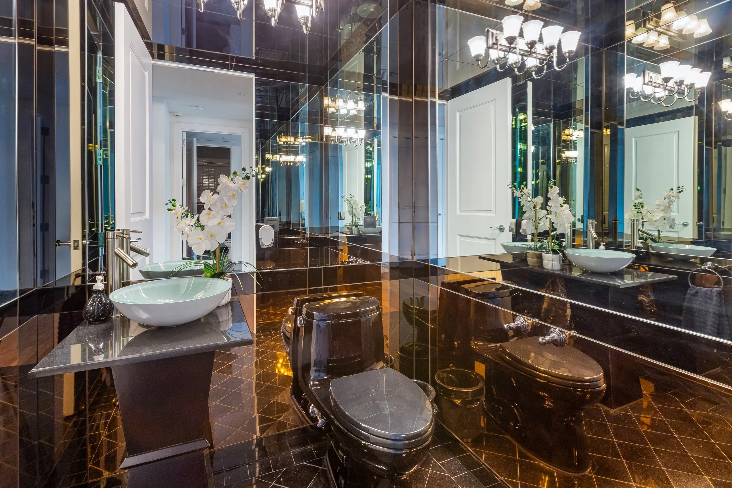 Luxurious bathroom featuring black tiled floors and walls, white vessel sinks with orchids, black toilets, mirrored walls, and modern lighting fixtures.