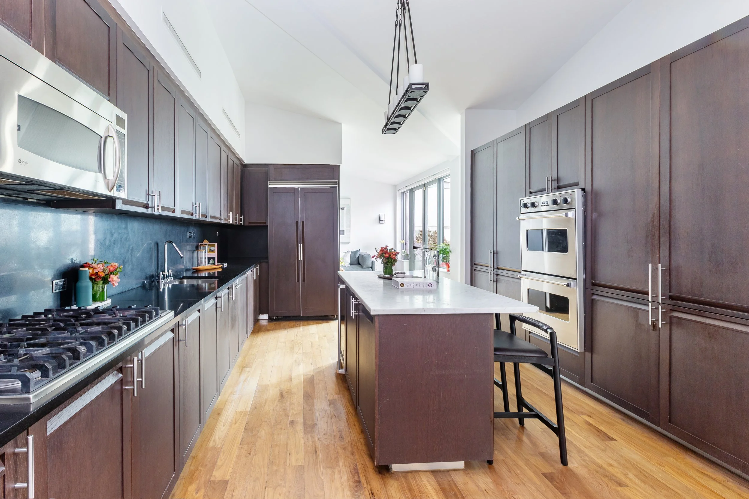 Modern kitchen with dark wood cabinets, white marble island, stainless steel appliances, and hardwood floors, with a view into a bright living area.