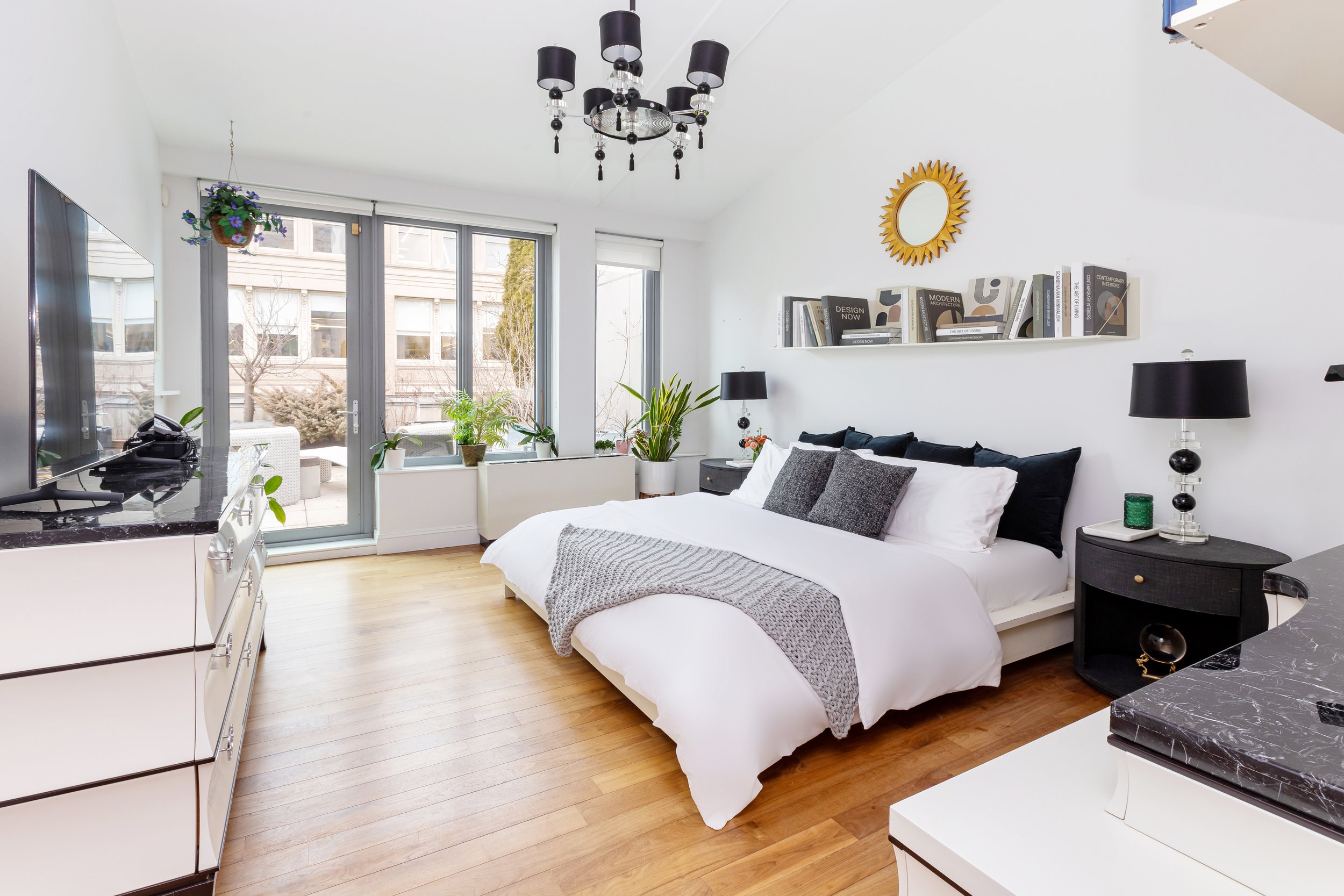Bright bedroom with large sliding glass door leading to outdoor patio, decorated with plants, featuring a white bed with black and gray pillows, black nightstands with black lamps, a black chandelier, and a white shelf with books above the bed.