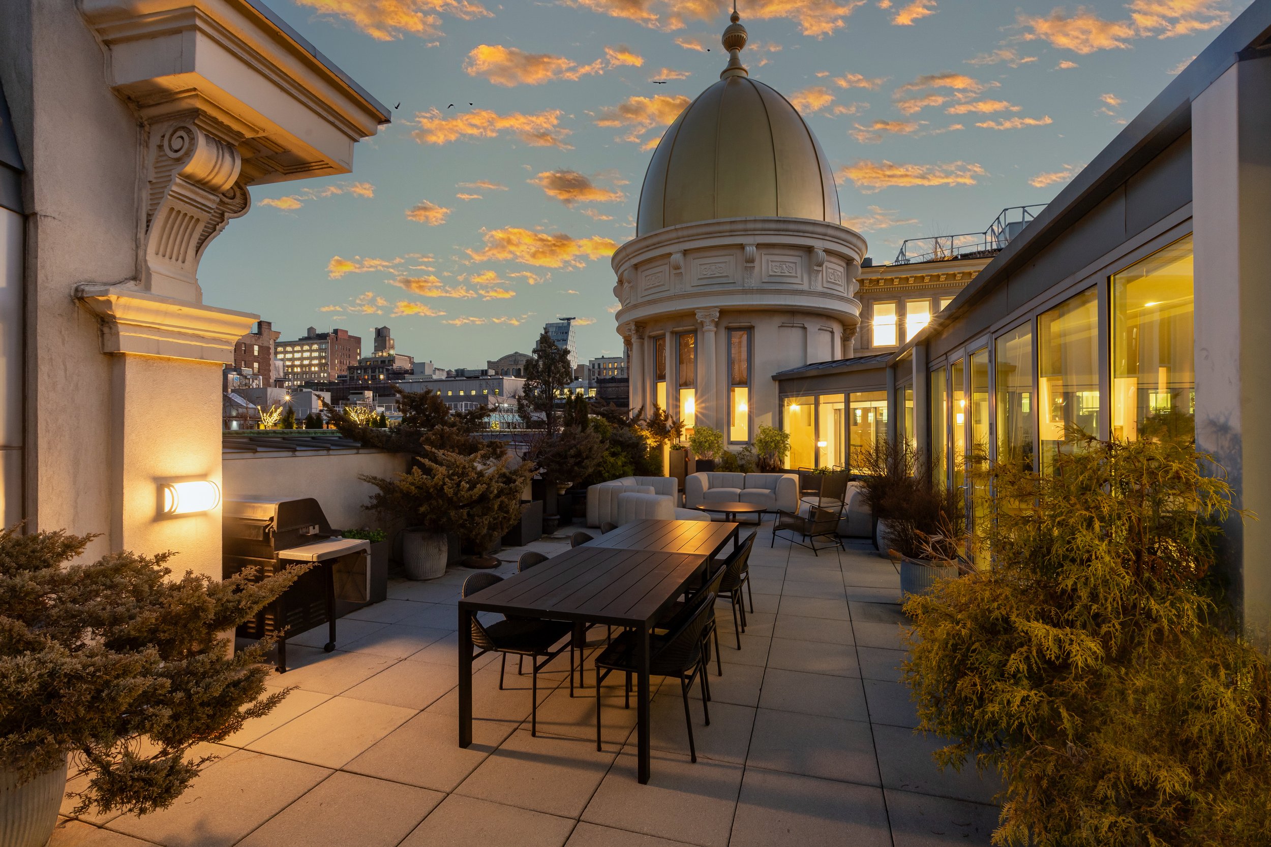 Rooftop terrace at sunset with outdoor seating, potted plants, and city skyline in the background.