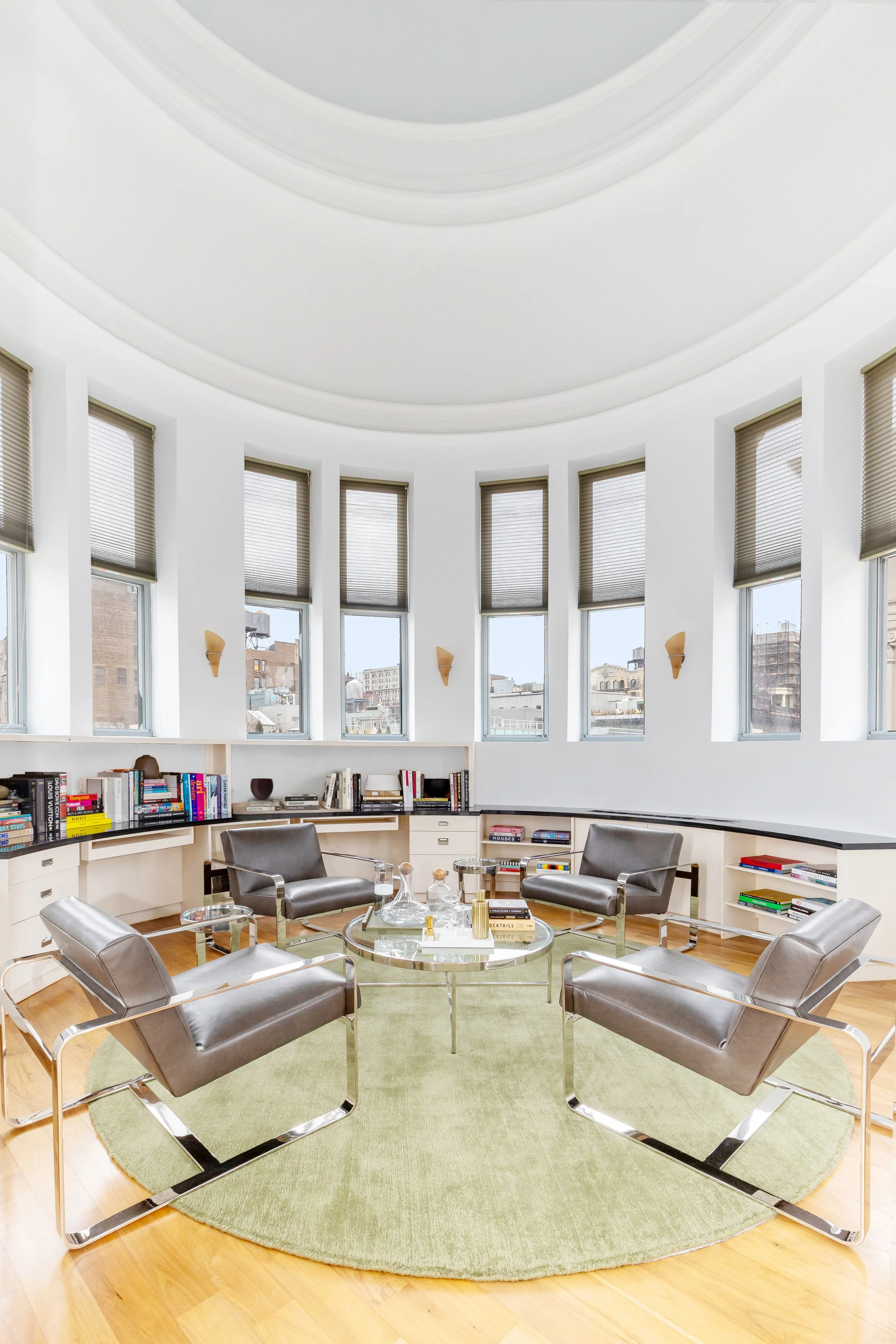 Modern meeting room with four gray chairs arranged around a glass coffee table on a round green rug, bookshelves along the perimeter with books and decorative items, large windows with gray blinds, and cityscape view outside.