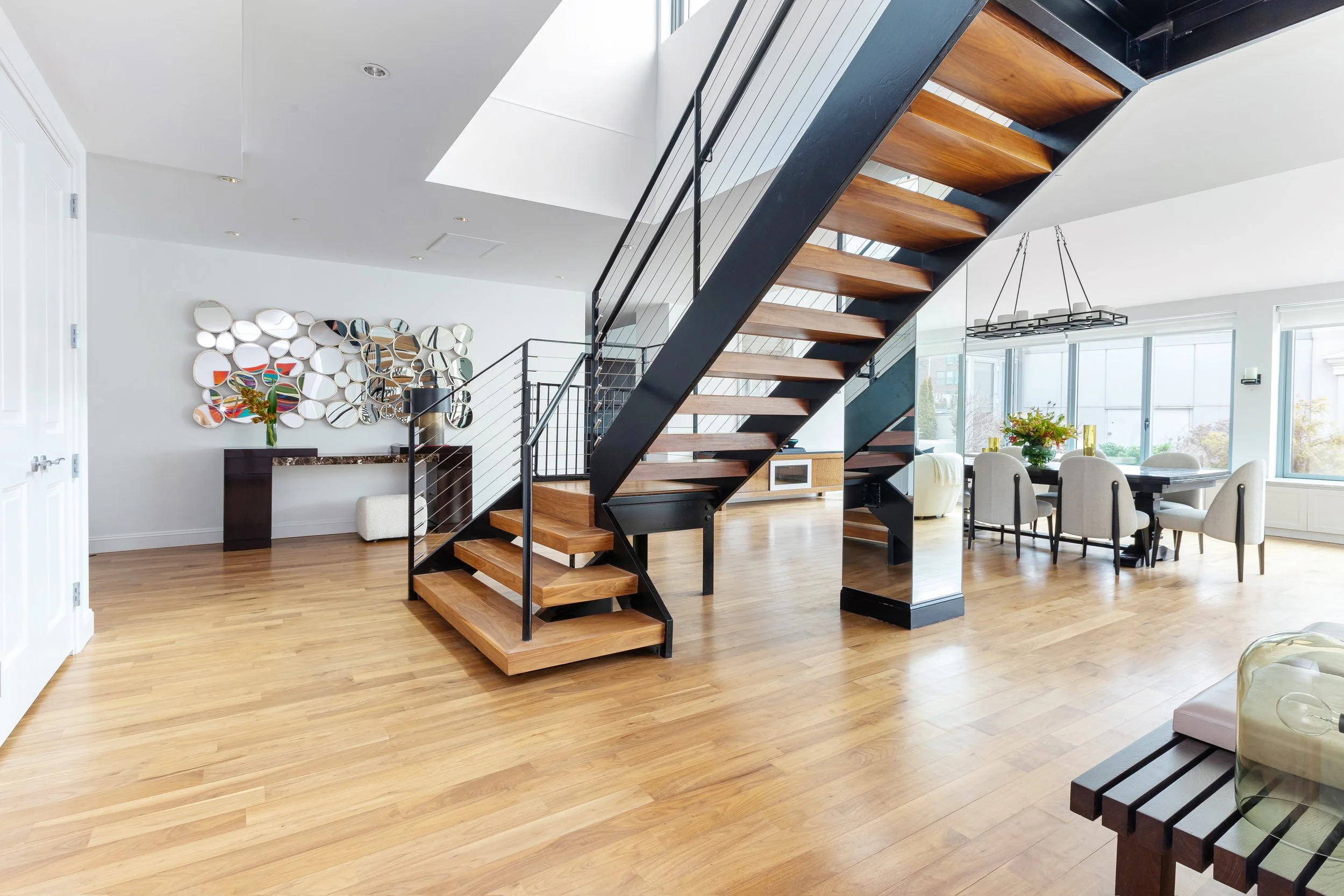 Interior of a modern home with a wooden staircase with black metal railing, dining area with six off-white chairs around a black table, large windows, and minimalistic decor.