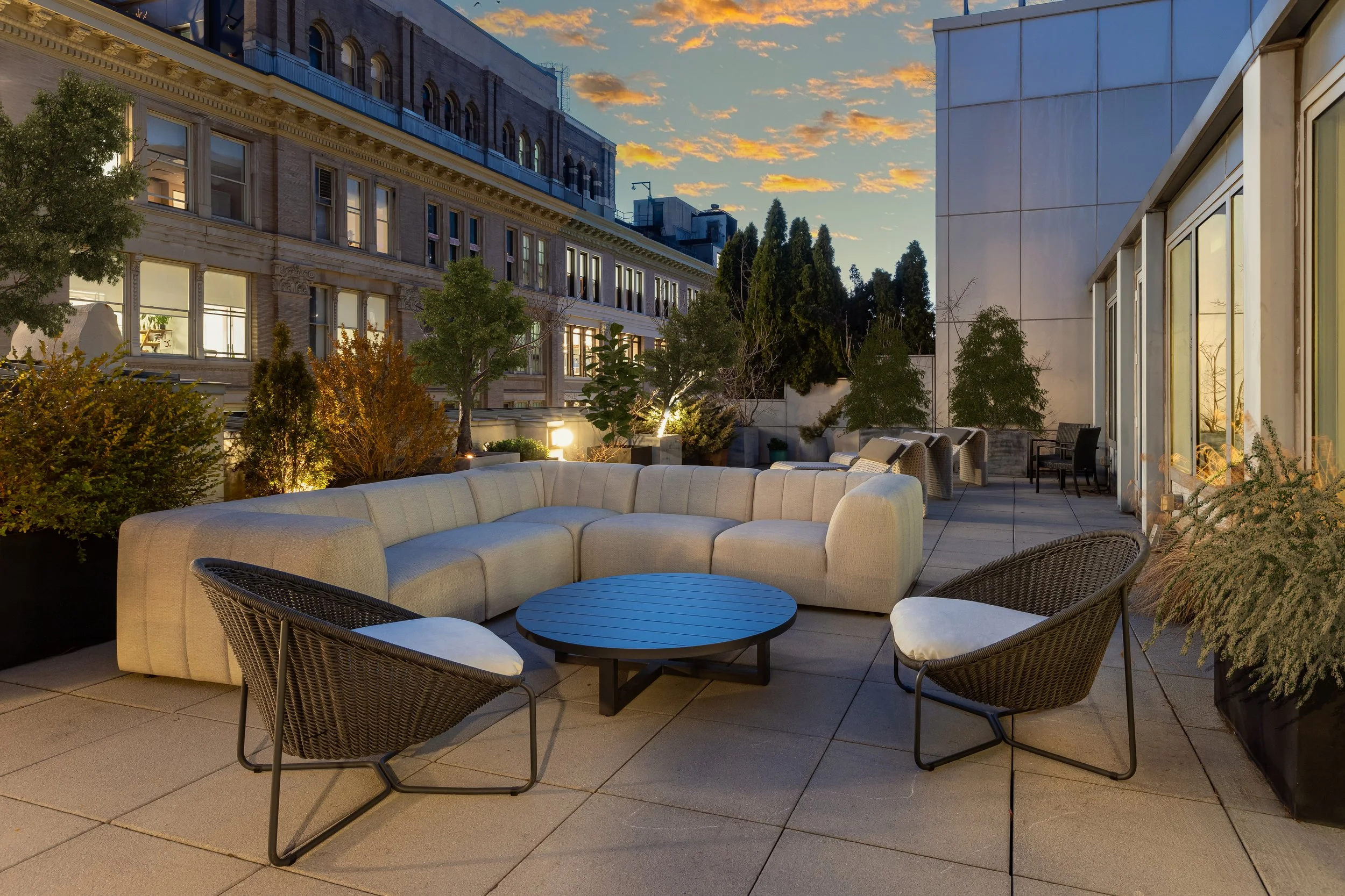 Outdoor rooftop patio with beige sectional sofa, black wicker chairs with white cushions, a round black table, and planters with greenery. Evening setting with a sunset sky and city buildings in the background.