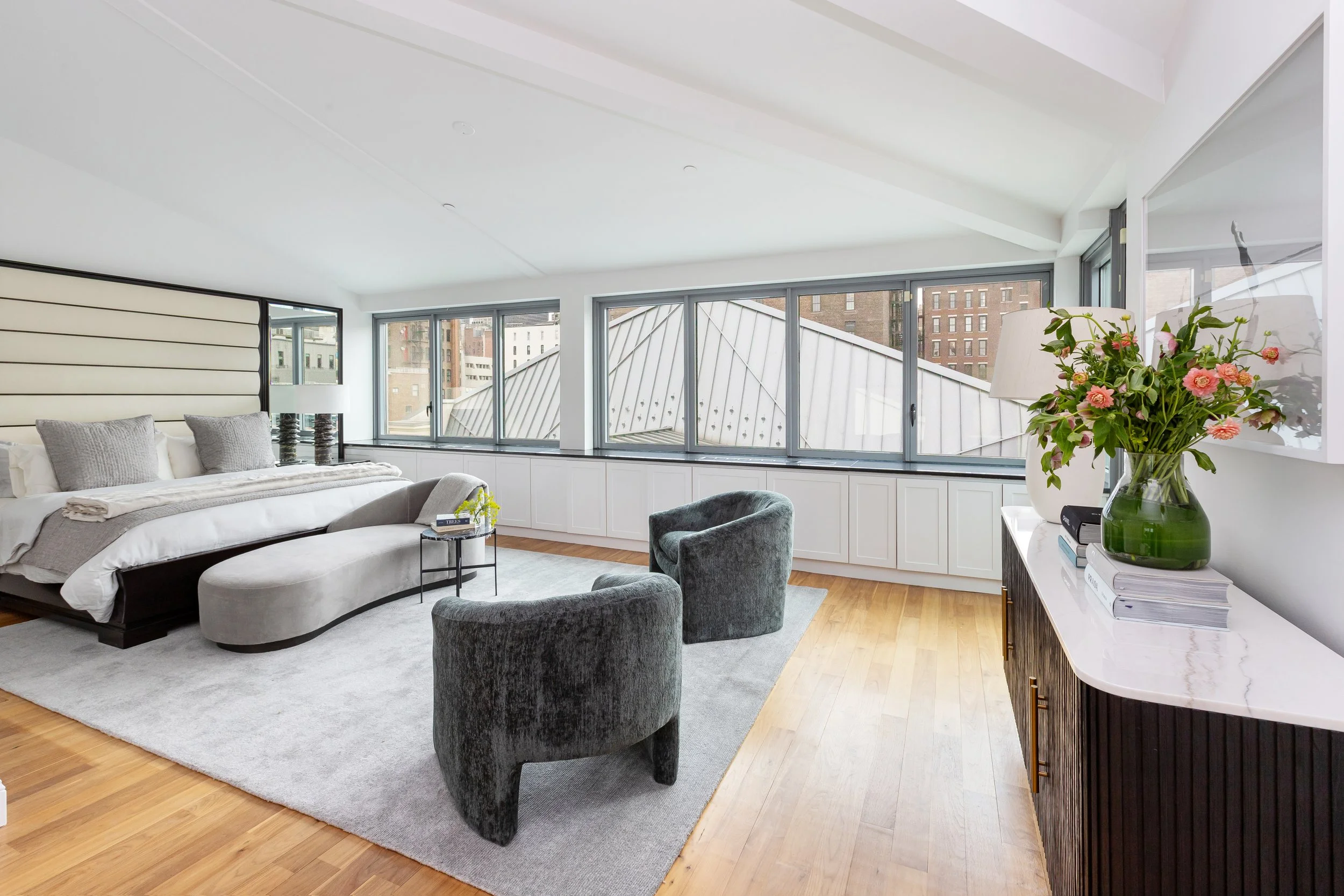 Modern bedroom with large windows, bed with gray bedding, black and gray chairs, a white rug, and a white sideboard with a vase of pink flowers.
