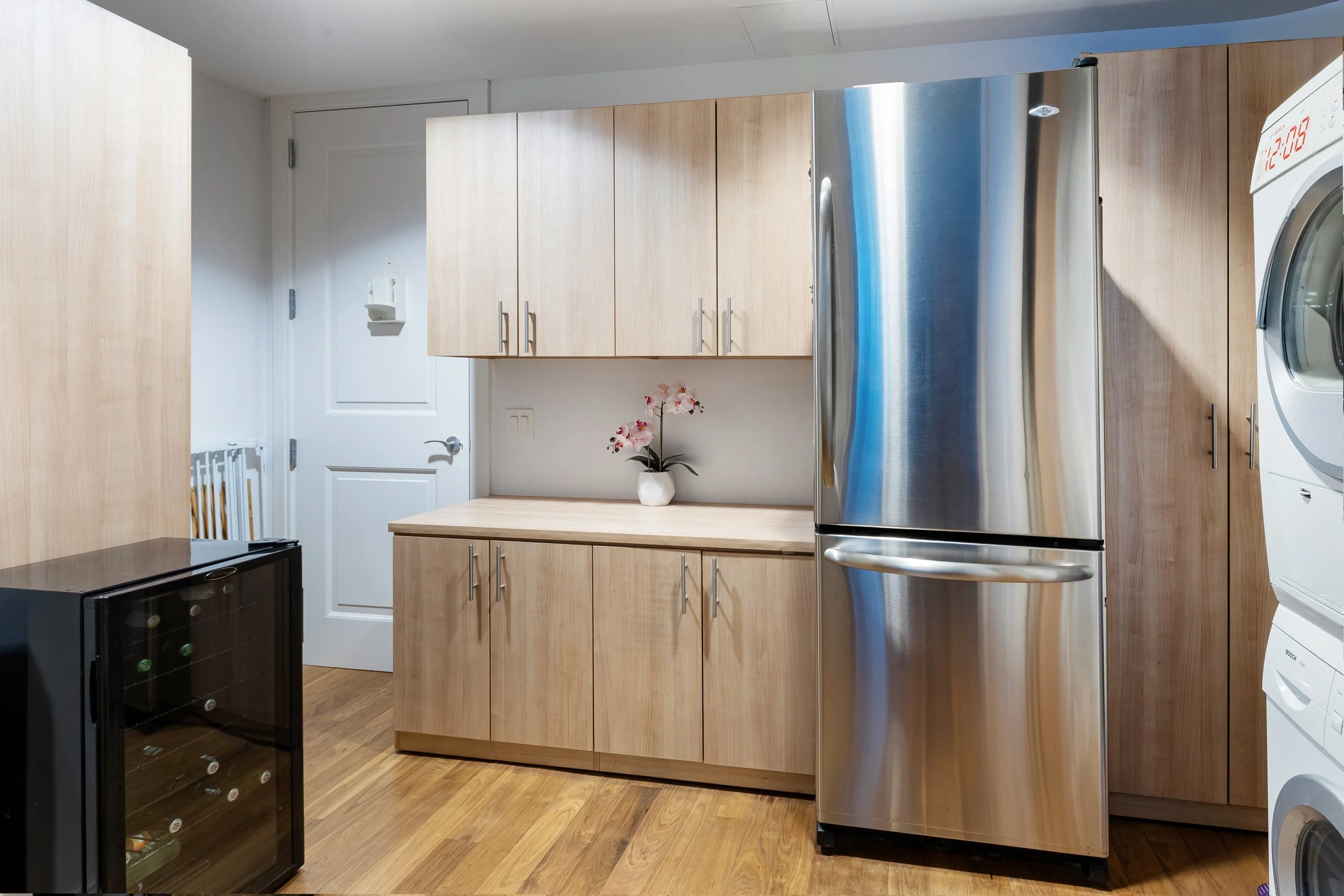 Kitchen with wooden cabinets, a stainless steel refrigerator, a small black wine cooler, a white door, a countertop with a potted pink orchid, and a stacked washer and dryer.