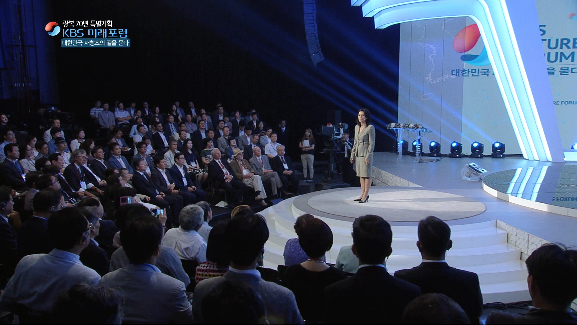 A woman in a gray business suit stands on a small round stage speaking at a conference, with an audience seated in front of her and behind her on tiered seating. The backdrop displays a large screen with a logo and text in Korean, and stage lighting illuminates the scene.
