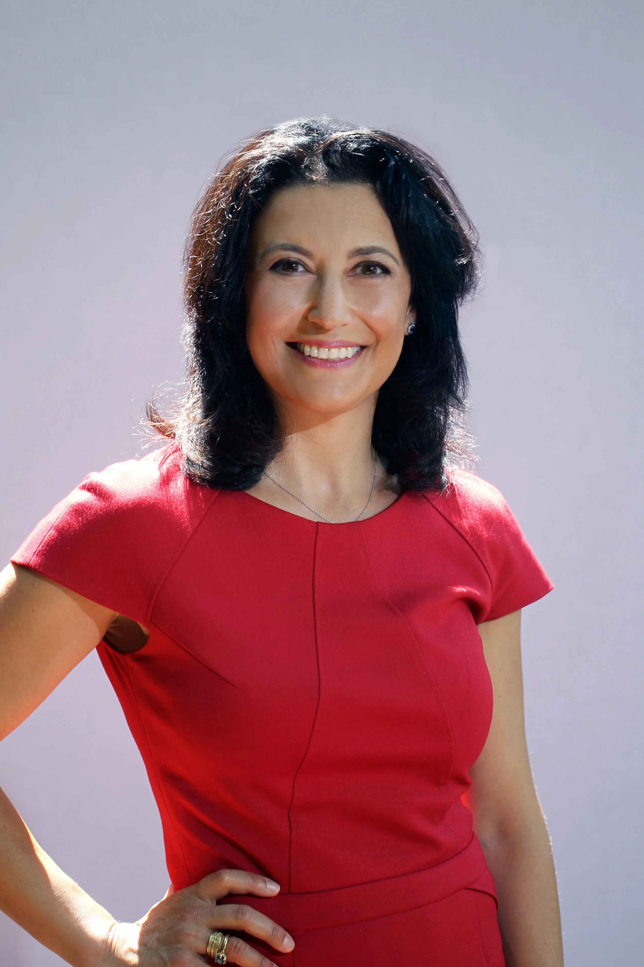 A woman with dark, shoulder-length hair, wearing a red dress, smiling and standing with one hand on her hip in front of a plain background.