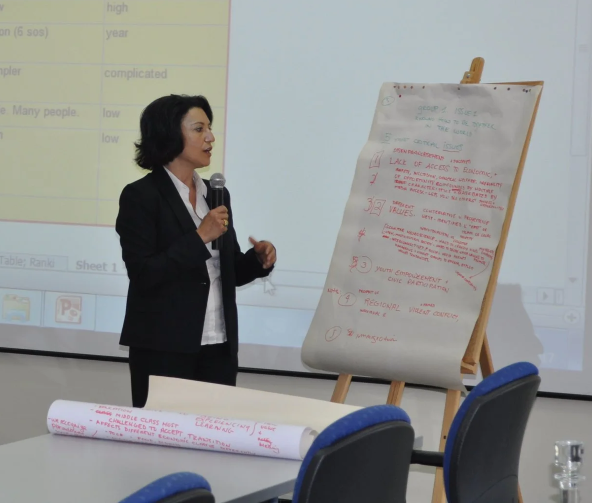 A woman in a black suit holding a microphone presents next to a large sheet of paper with handwritten notes on a stand in a conference room. There is a projection screen in the background with a slide displaying a list.