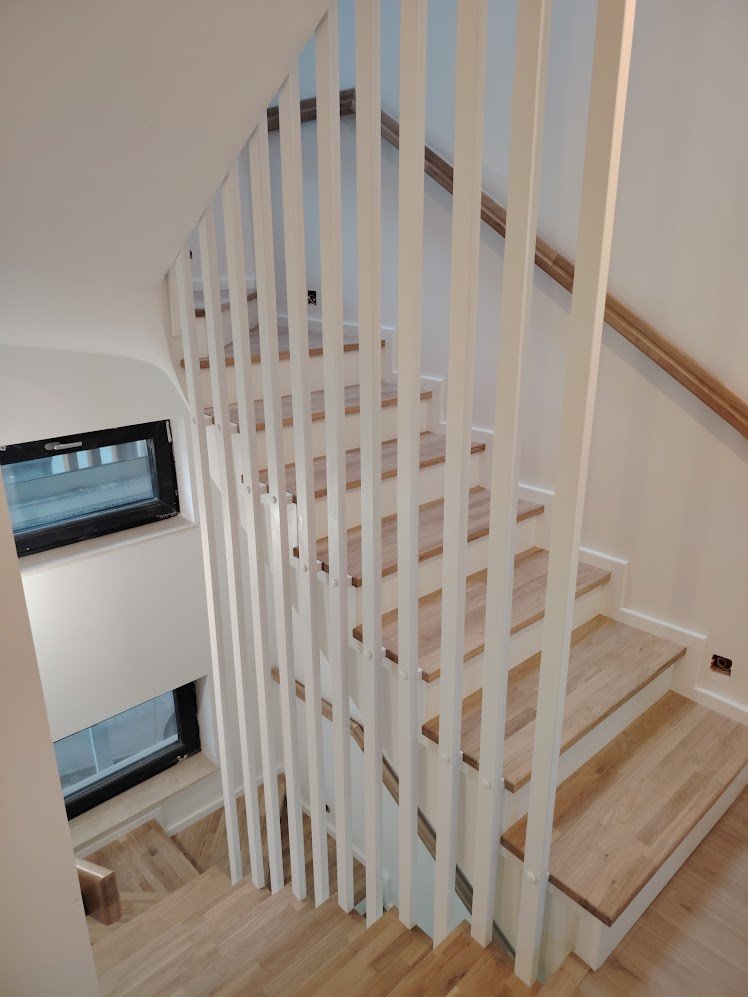 Indoor staircase with wooden steps and white vertical slats, featuring a wooden handrail along the wall, and a small window near the bottom of the stairs.