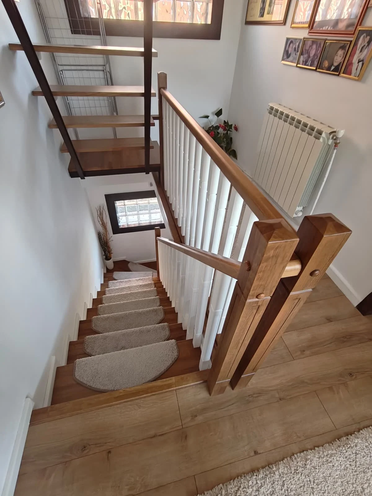 View of a staircase from the top, showing carpeted steps, a wooden handrail, and a wall decorated with framed photos. There is a window at the landing and a radiator on the right wall.