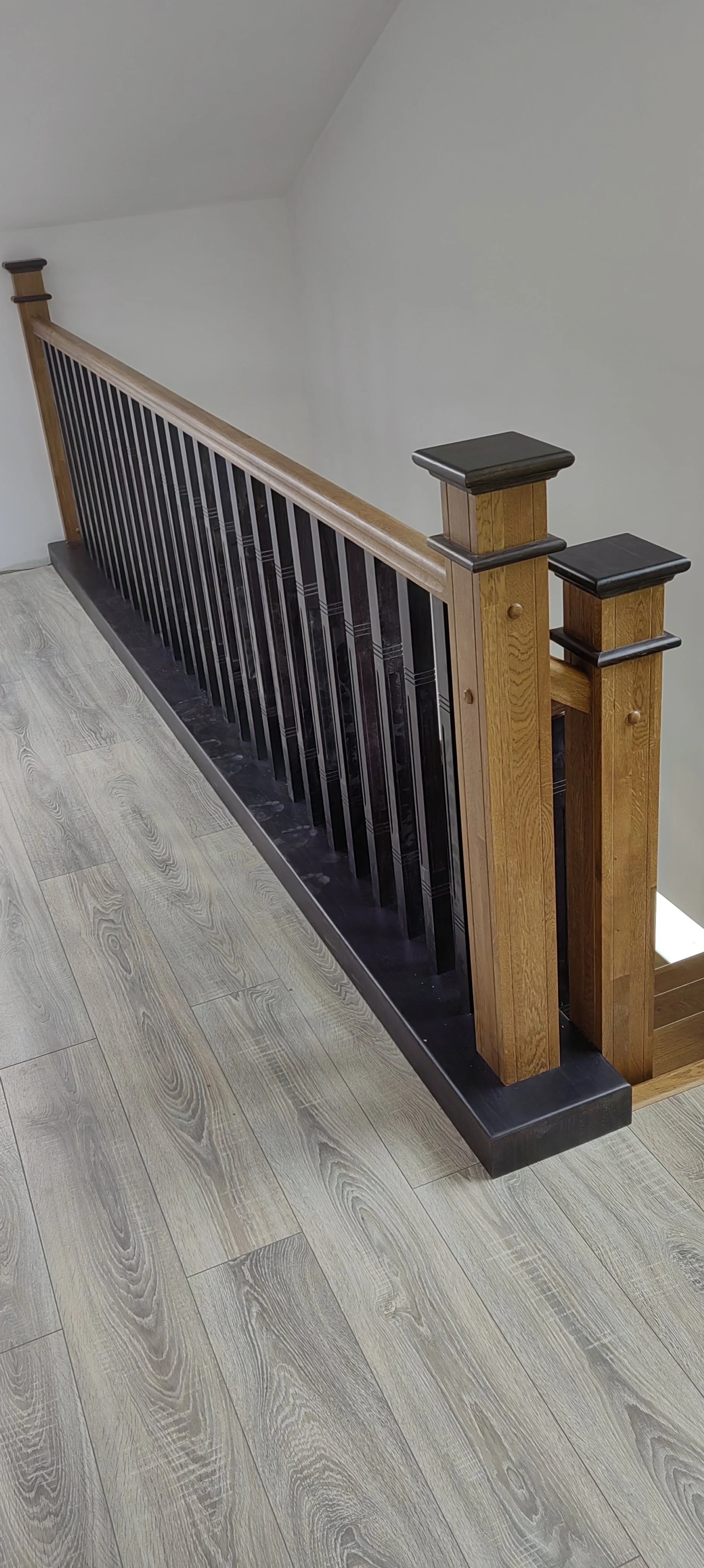 Wooden staircase banister with black metal bars, light wood posts with black caps, and a black base at the bottom, next to light wood flooring and a light-colored wall.