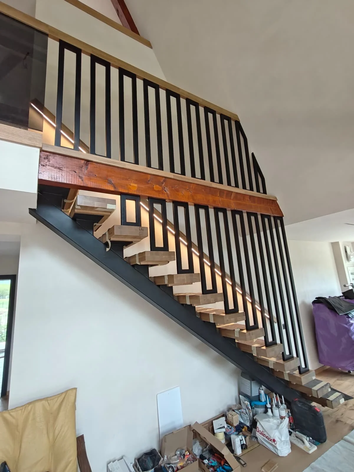 Interior view of a modern staircase with wooden steps, black metal railing with vertical bars, and a wooden handrail, leading to an upper floor in a house under construction or renovation.