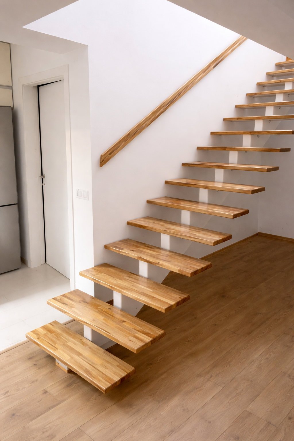 Interior shot of a modern wooden floating staircase with a wooden handrail, adjacent to a white wall and a partial view of a kitchen with stainless steel appliances.