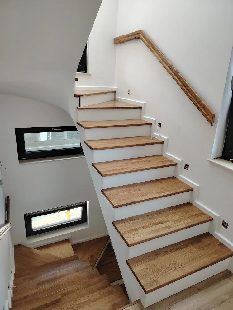 Wooden staircase with a white base and black handrail, leading upstairs in a modern home with white walls and windows.