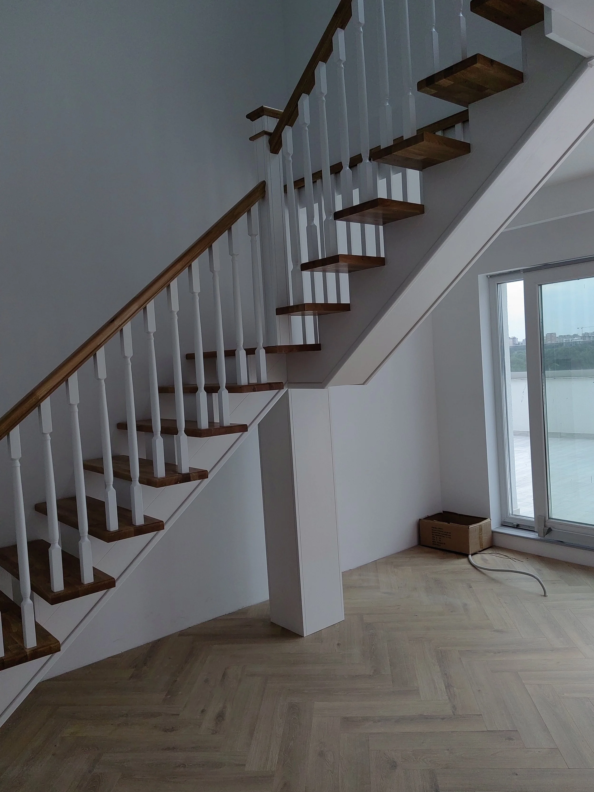 Interior view of a recently renovated staircase with wooden steps and white balusters, leading to an upper floor, next to a large glass sliding door showing a view outside.