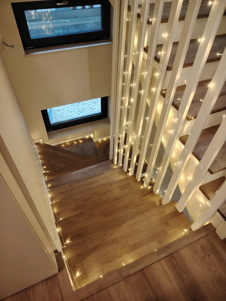 Wooden staircase decorated with warm white string lights, seen from the top looking down, with two rectangular windows along the stairway wall revealing an outdoor scene.