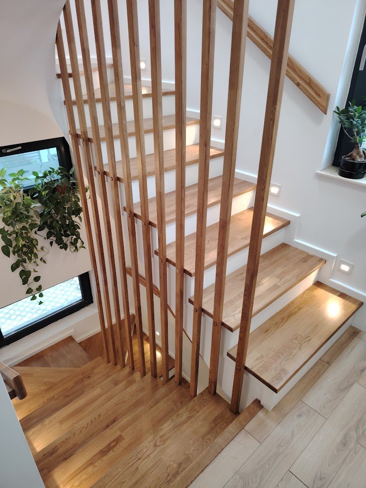 Wooden staircase with integrated lighting, viewed from above, with a railing made of vertical wooden slats and a window with plants nearby.