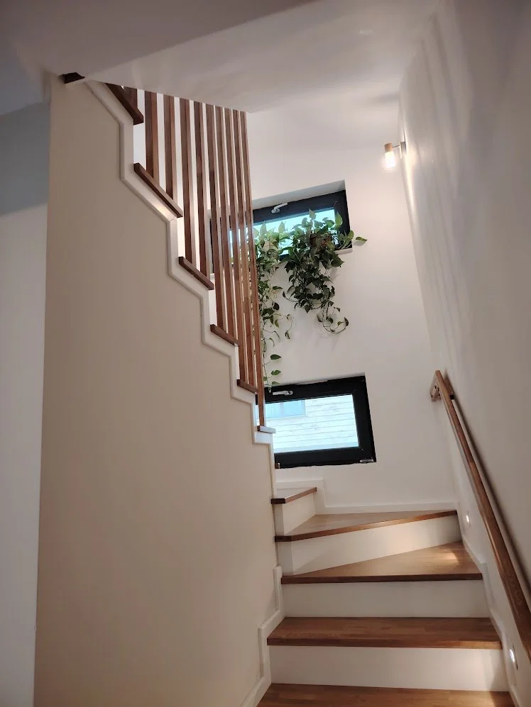 Interior view of a staircase with wooden steps, white walls, a wooden handrail, two windows, and a potted plant near the top window.