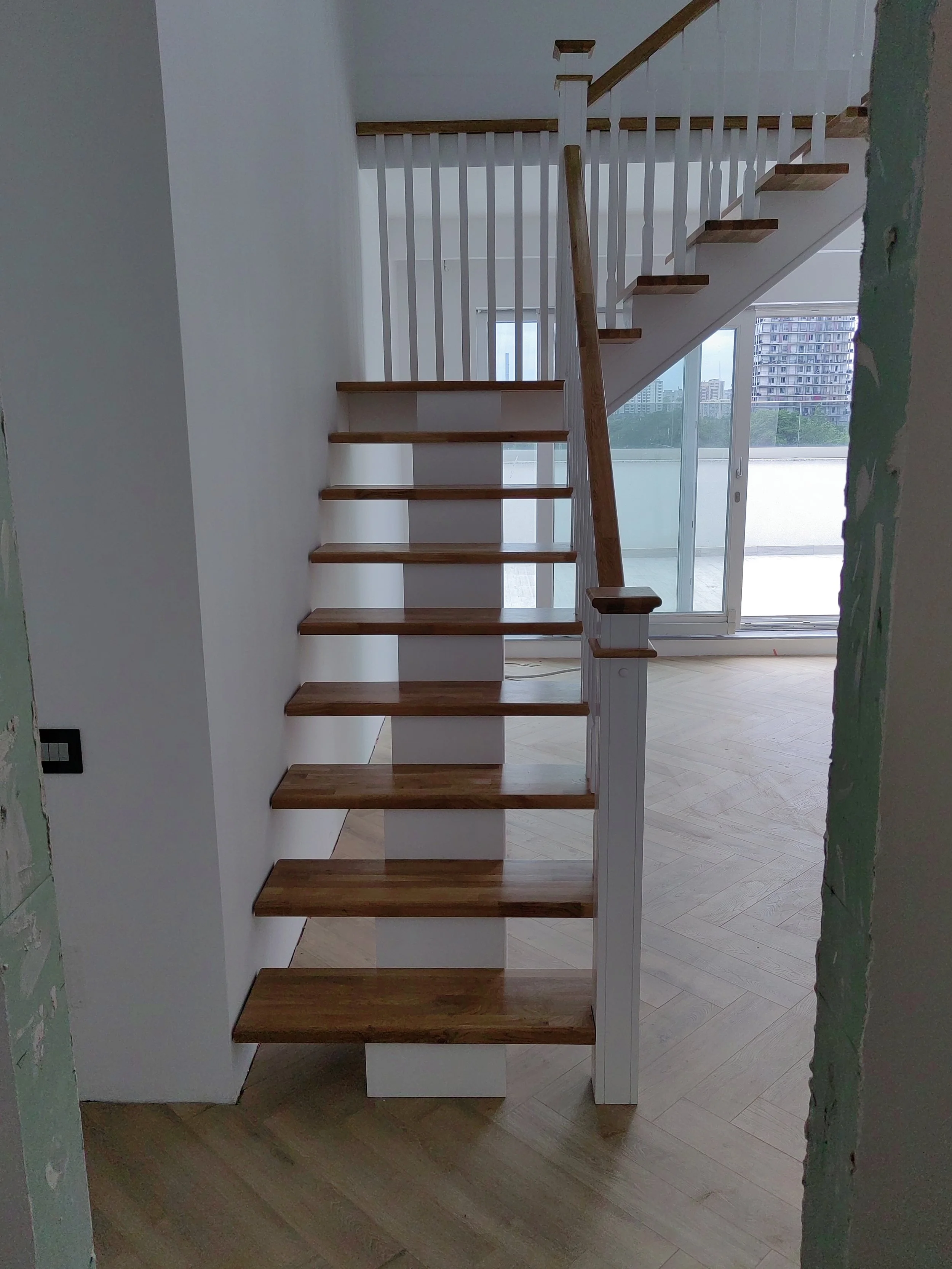 Photo of an interior staircase with wooden steps and white railings, leading to an upper floor. Visible windows in the background show city buildings.