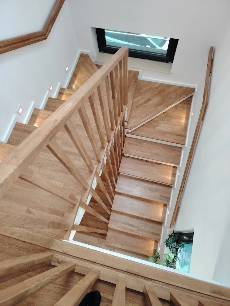 Angled view of a wooden staircase with a handrail, leading down to a lower level with light-colored walls, small square wall lights, and a window with plants nearby.