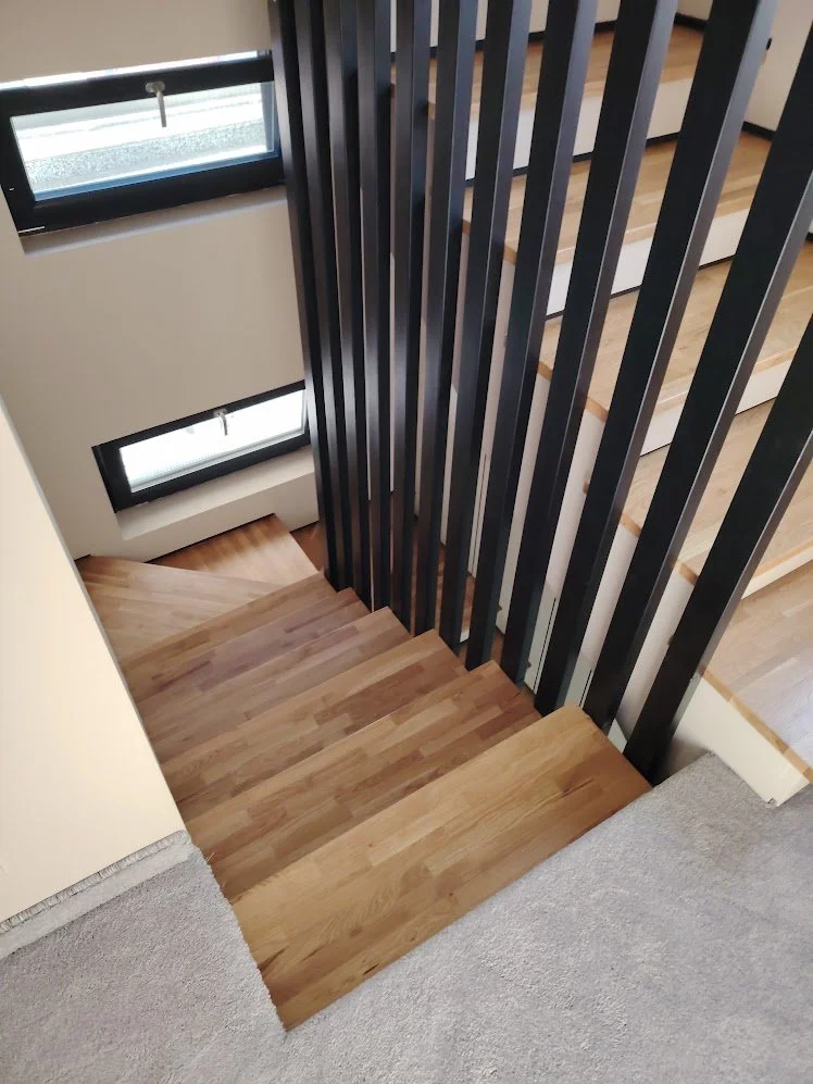 Wooden staircase leading downward with a black vertical railing, adjacent to large windows with horizontal blinds, in a modern interior.