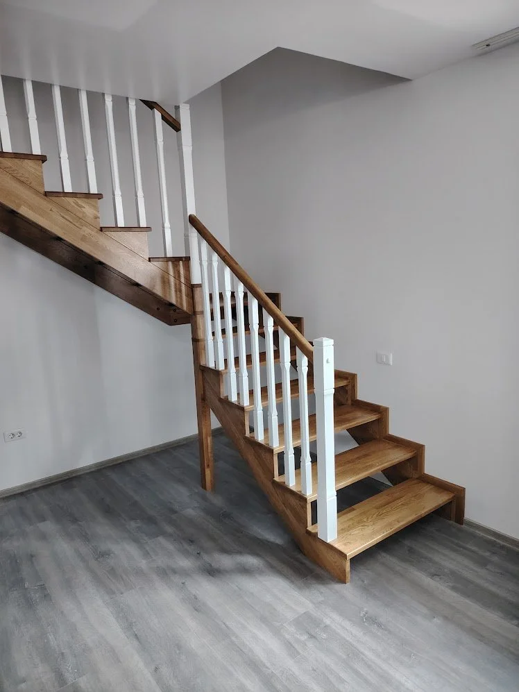Interior view of a wooden staircase with white spindles and a wood handrail, leading to an upper floor, in a room with gray flooring and white walls.