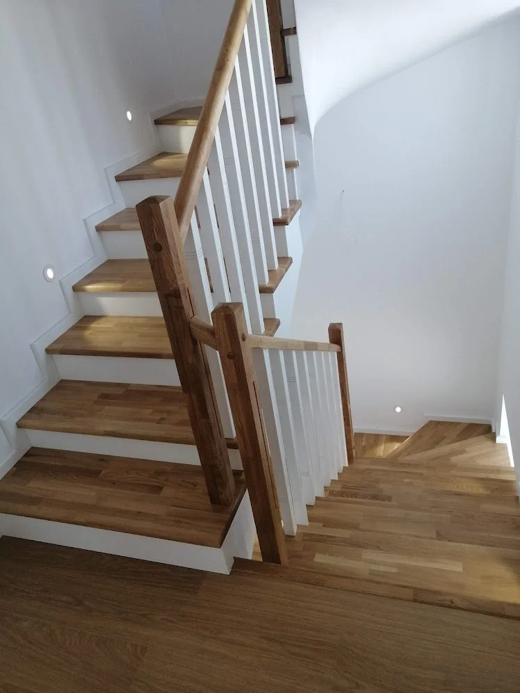 Interior view of a wooden staircase with white risers and handrails, leading upstairs and downstairs, in a well-lit area of a house.