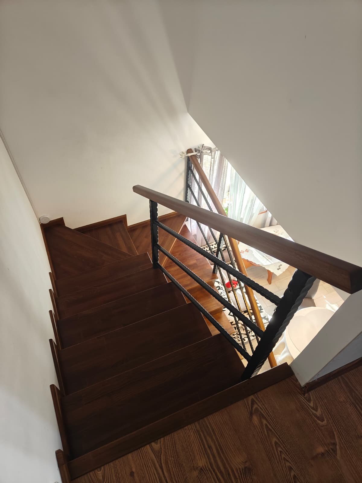 Wooden staircase with black metal railing leading down to a room with a sofa and a small table near a window with curtains.