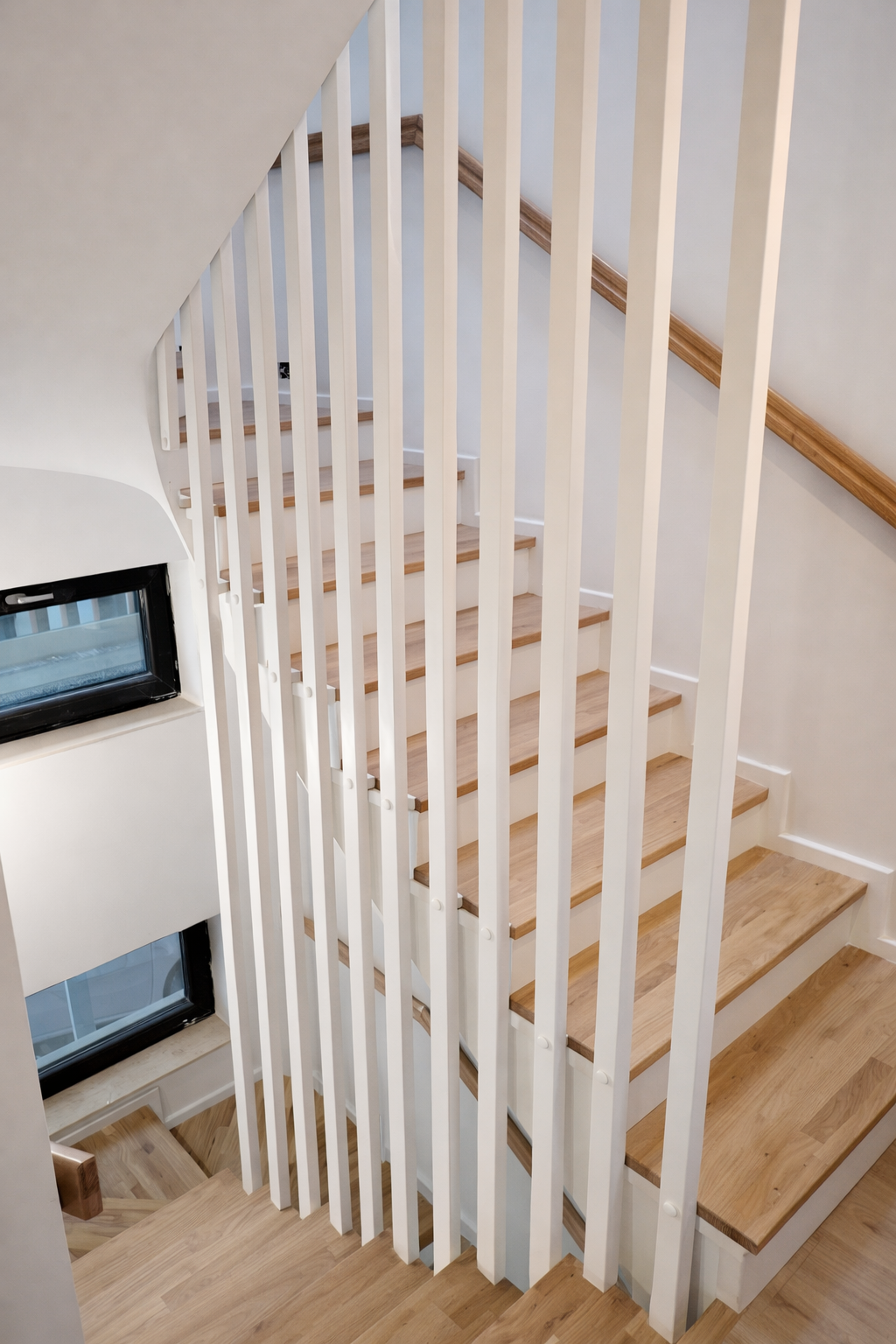 Wooden staircase with white vertical spindles and wooden handrail, next to a wall with a small window.