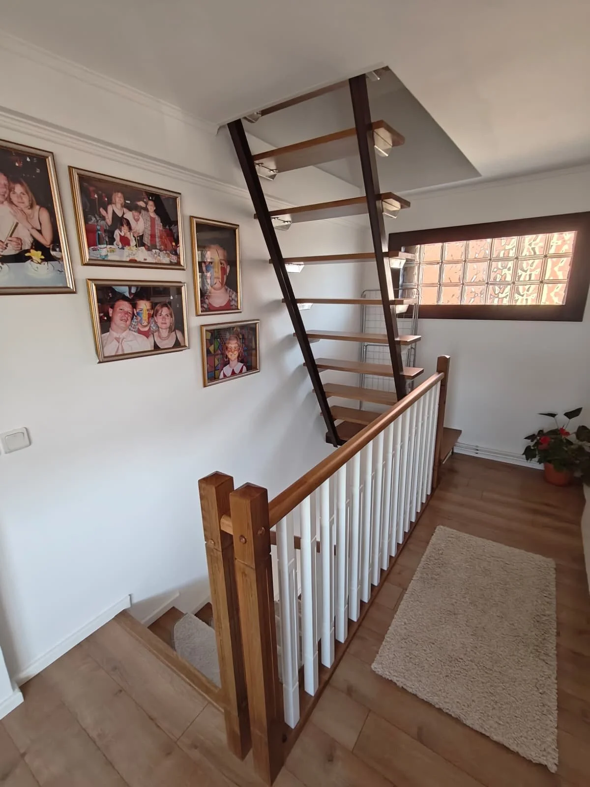 Interior view of a staircase landing with framed family photos on the wall, a wooden handrail, a potted plant, and a window with decorative brickwork.