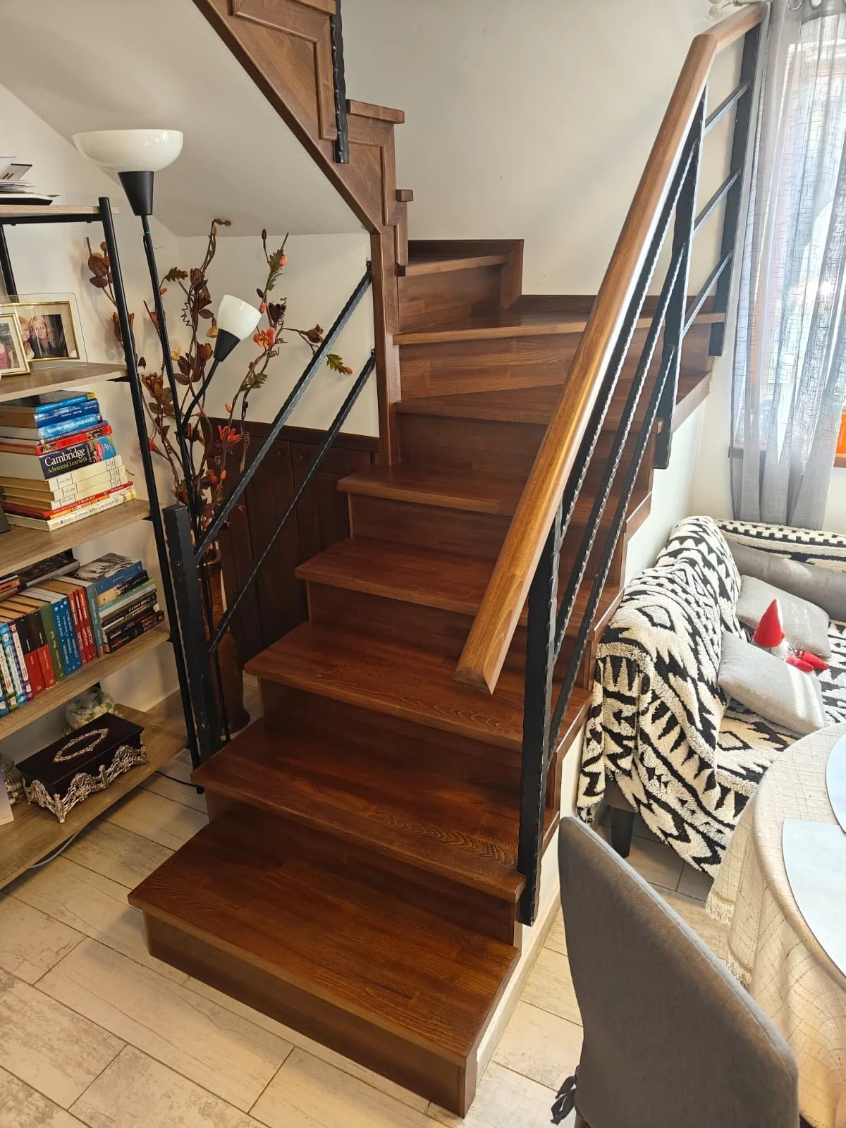 Photo of a wooden staircase with black metal railing in a living room. To the left, a bookshelf filled with books, and a tall decorative lamp with multiple branches and white lampshades. To the right, a patterned sofa with a blanket, pillows, and a w