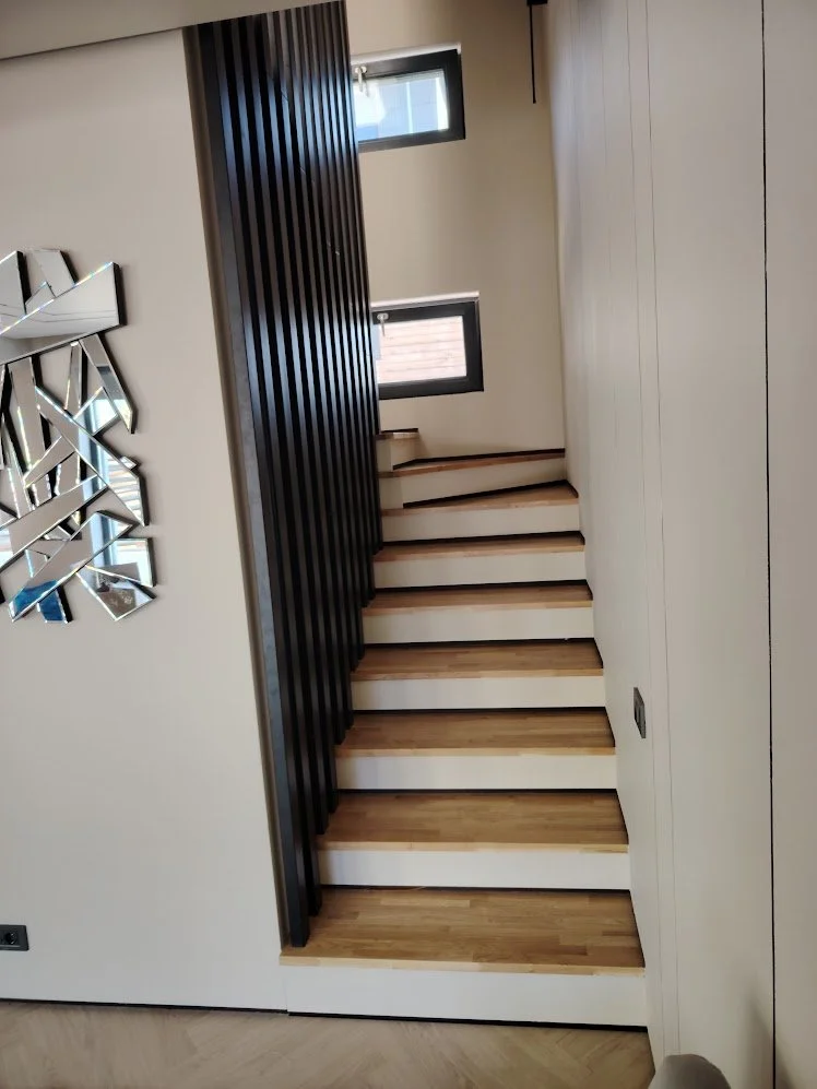 Interior view of a staircase with wooden steps, black vertical railing, two small windows, and a decorative mirror on the wall.
