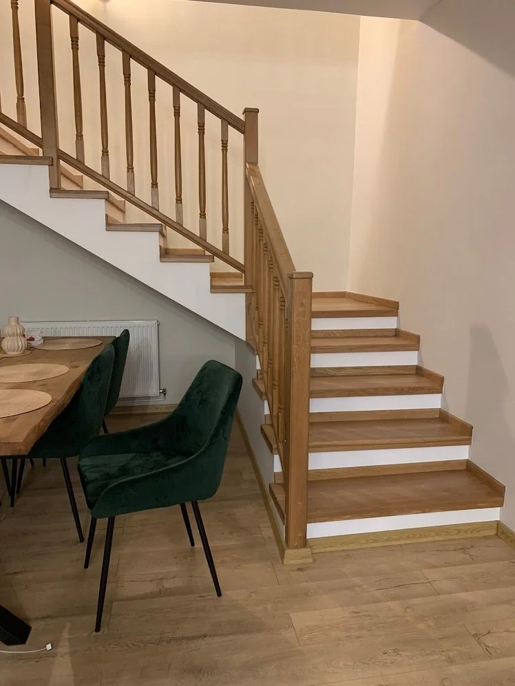 Wooden staircase with vertical spindles leading upstairs inside a house, next to a dining area with a table and green chairs.
