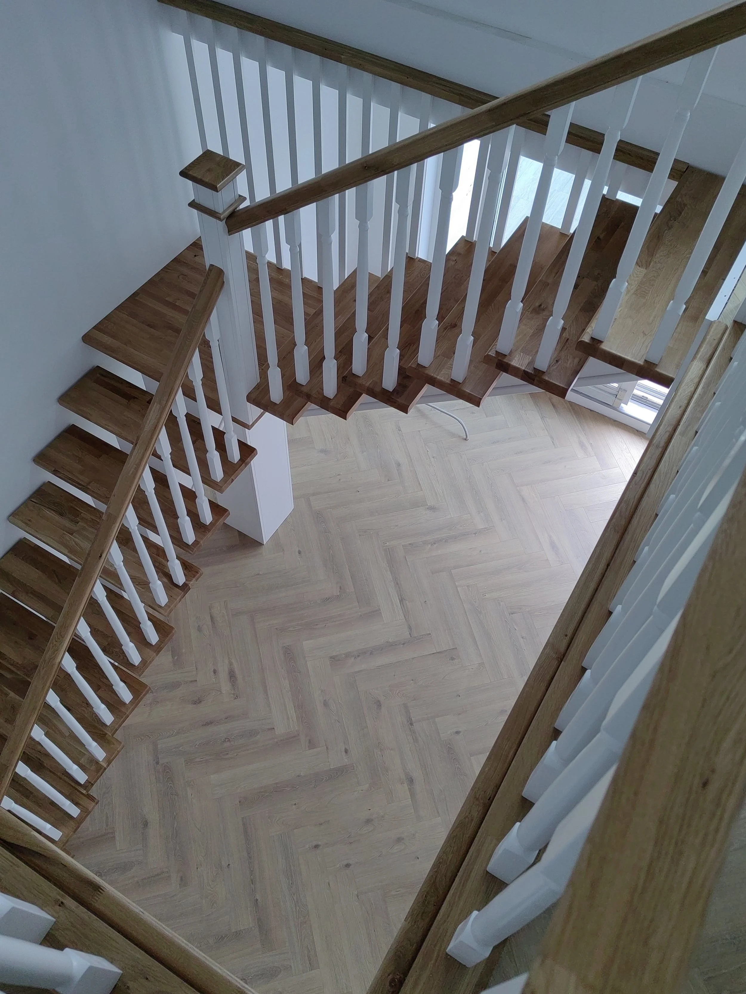 Inside view of a staircase with wooden handrails and white balusters, overlooking a room with wooden flooring and a window letting in natural light.