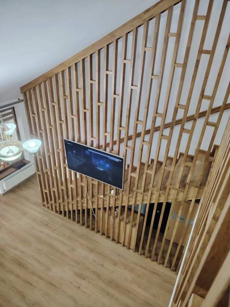 Interior view of a wooden staircase surrounded by a wooden slat partition, with a TV mounted on the wall and a chandelier with glass shades visible in the corner.