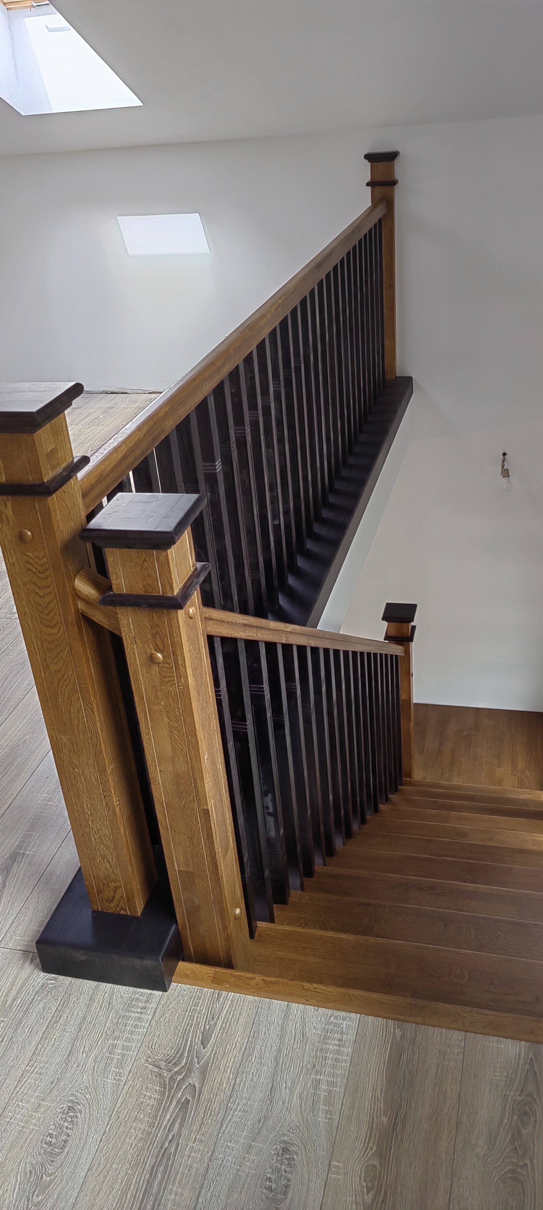 Wooden staircase with black rails and newel posts leading down, with an open space with white walls and skylights.