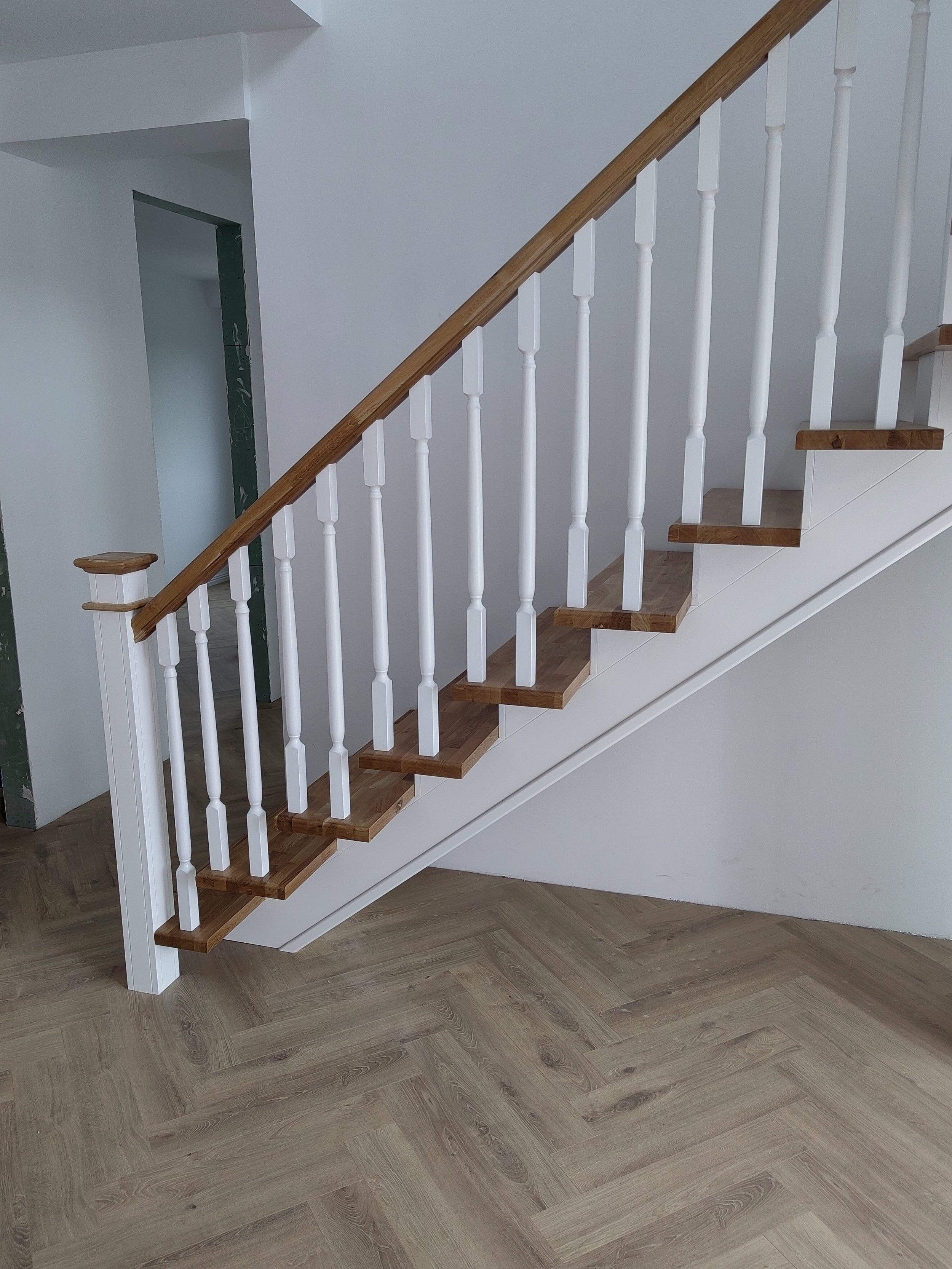Interior view of a staircase with wooden steps, white painted spindles, and a wooden handrail, with light hardwood flooring and plain white walls.