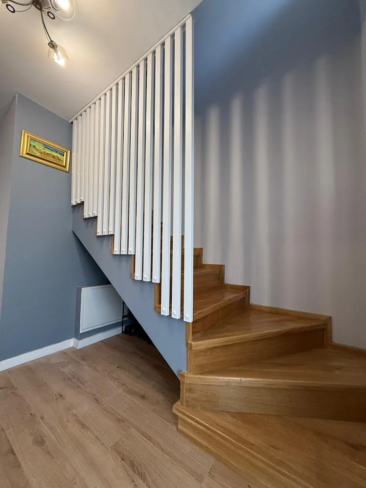 Interior view of a staircase with wooden steps and a white railing, against light blue walls, with a framed landscape painting on the wall and ceiling lighting fixture.