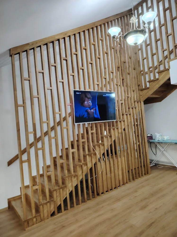 Wooden staircase with a decorative railing and a mounted flat-screen TV on the wall below; a chandelier hanging from the ceiling.