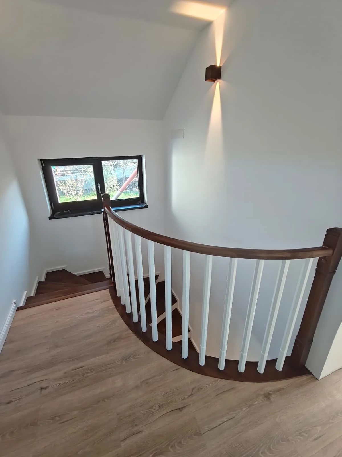 Interior view of a staircase with wooden steps and white balusters, a wooden handrail, a small window, and a wall-mounted light fixture.