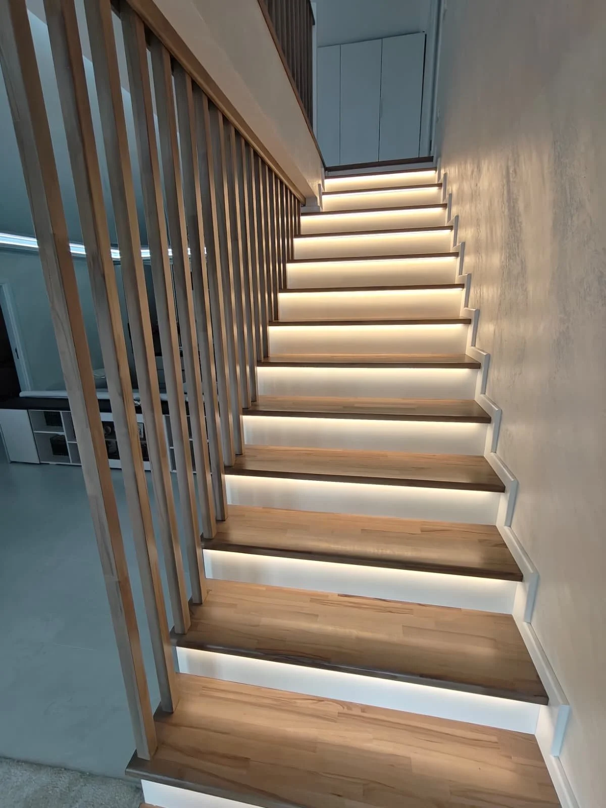 Modern staircase with wooden steps and LED lighting underneath each step, railing made of vertical wooden slats, leading to an upper floor in a contemporary home.