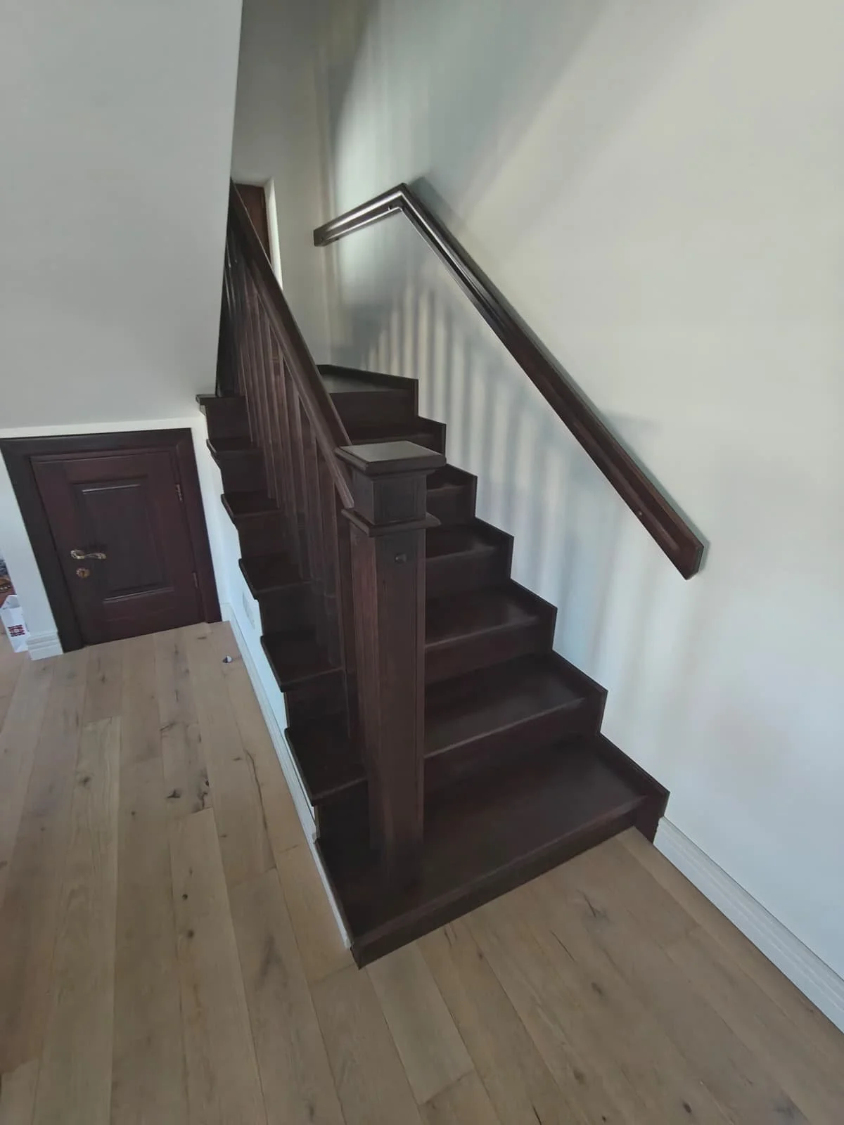 Dark wood staircase with a matching handrail and a small door underneath, adjacent to a light-colored wall and light wooden flooring.
